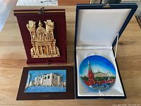 Front view of three decorative items: golden Petra plaque, a blue round Moscow plate in box, and a framed Tripoli relief.