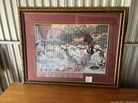 Framed print artwork showing an outdoor dinner scene at the Ritz Hotel Paris with people dressed in period attire, detailed garden and string lights, in a gold-tone frame with burgundy and cream mats.