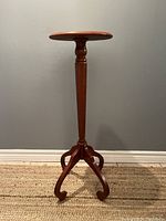 Side view of the mahogany plant stand showing the round top and tripod curved legs on a beige woven rug with gray wall background.