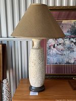 Front view of mid century modern textured ceramic table lamp with shade on top, sitting on wooden surface in front of framed painting.