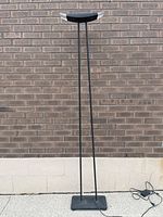 Full view of the tall postmodern floor lamp standing against a brick wall, showing the slim metal base and vertical rods supporting the upper crescent-shaped lamp head with acrylic ends.