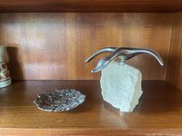 Wide view showing the aluminum goose sculpture on granite base and the floral pin dish beside it on a wooden shelf.