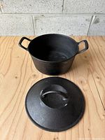 Open cast iron pot and separate lid on wooden surface with cinder block wall background.