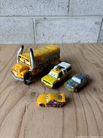 Four Disney Pixar Cars toy cars on wooden surface, showing Mack truck and smaller character cars.