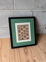 Framed textile artwork with green matte displayed on wood surface against a concrete block wall