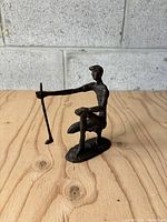 Front side view of the MCM brutalist golfer sculpture figure, showing the golfer holding the golf club upright, standing on a wooden surface with a concrete wall background.