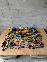 Overview photo of all the toys spread out on a wooden table including various toy cars, trucks, action figures, and a toy airplane.