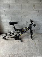 Black Mongoose RX2.6 mountain bike frame standing against a concrete block wall, showing overall frame with seat, pedals, and partial drivetrain.