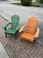 Pair of green and orange wooden Muskoka chairs pictured outdoors on paved and concrete surface.