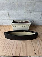 Front view of black glazed canoe-shaped planter alongside off-white rectangular planter on wooden surface against grey brick wall.