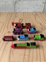 Seven toy trains from Thomas the Tank Engine and Friends arranged on a wooden surface against a concrete block wall, showing front and side views.