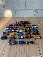 Lot of 33 toy cars arranged in a grid on a wooden surface, viewed from above, showing various colors and models.