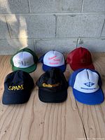 Six advertising hats including SPAM, Continental, Primetel Evans Margate, Reuland Electric, PCL, and a red solid cap photographed on a wooden surface in front of a concrete block wall.