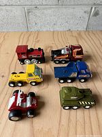 Top-down view of six vintage toy trucks made by Buddy L, showing the red locomotive, blue dump truck, red and yellow semi, yellow Challenger truck, white and red tractor, and green tank on a wooden surface.