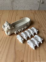 Main image showing the large Fitz And Floyd pig cracker tray and eight smaller pig napkin holders arranged next to it on a wooden surface.