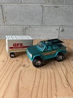 Green Nylint Chevy Tahoe toy truck with gold Tahoe decals and black roof, next to white Gordon Food Services box trailer with red text on wooden surface.