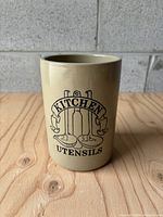 Front view of the beige stoneware kitchen utensil container showing the 'Kitchen Utensils' artwork and textured wood surface background.