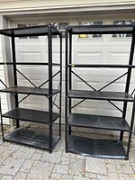 Two black metal shelving units side by side outdoors on a paved surface against a garage door background. Each unit has four shelves and a metal frame with cross braces.