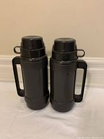 Two black thermoses side by side showing front profiles with handles and cup lids attached.