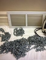Photo showing multiple metal chains coiled loosely on a surface next to a white rectangular cold air vent cover with horizontal slats.