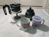Photo showing coffee percolator, tea infuser pot, two floral cups including one with lid, placed on white cloth