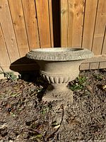 Side view of the concrete urn placed outdoors against a wooden fence, showing its shape, decorative details, and weathered surface.