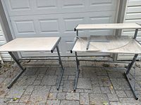 Photo of two rectangular desk tables with light wood grain surfaces and gray metal bases placed outdoors on pavement.