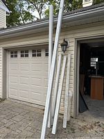 Metal and wooden downspouts leaning against garage and house exterior, showing condition and quantity.