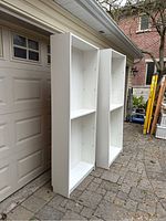 Two white IKEA Billy shelving units standing outside by garage door, visible adjustable shelf holes inside