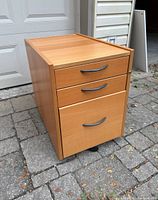 Front view of the IKEA beech finish drawer unit showing three drawers with silver handles on wheels, placed on stone pavement.