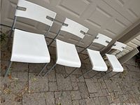 Four white plastic and grey metal IKEA stacking chairs arranged in a line on outdoor brick paving. Shows front and side views highlighting seats and backs. Condition scratches and marks visible.