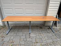 Full view of two IKEA Galant tables joined together, wooden beech colored plank tops, silver metal adjustable legs, placed outdoors on stone floor.