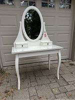 Front angled view of white vanity or desk with oval mirror and two small drawers attached above one large drawer and curved leg base.