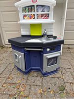 Frontal view of the plastic Little Tikes play kitchen showing various components including the stove burners, sink with faucet, oven with clear door, pantry cabinet, and a small upper window with play items inside.