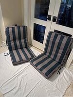 Two striped patio chair cushions with attached seat and back portions and ties for securing, arranged in seated position indoors.