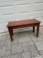 Full view of solid oak piano bench showing front and side, placed outdoor on stone pavement in front of a garage door.