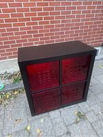 Wood grain laminate shelf unit with four red plastic drawers visible from front, outdoor setting.