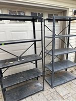 Two black metal shelving units side-by-side outdoors in front of a garage door. Both have five shelves with visible rust patches and scratches.