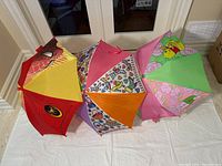 Three children's umbrellas displayed fully opened showing designs: Spider-Man red and yellow, colorful girl pattern, and Winnie the Pooh pink and green.