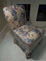 Front angle view of vintage small upholstered chair showing curved backrest and skirted bottom with floral fabric pattern wrapped in clear plastic.