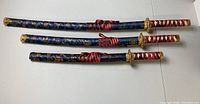 Set of three decorative Japanese-style swords lying flat on white surface: large, medium, and small sizes with blue sheaths and red and white wrapped handles.