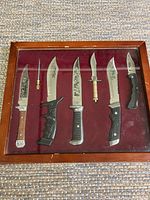 Full view of all 6 Bowie knives and sharpening rod displayed in wooden framed glass case with burgundy fabric backing. Scratches visible on wood frame, knives show decorative blade etching.