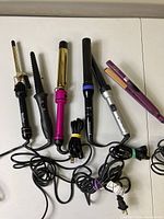 Five curling irons and one purple hair straightener laid out with cords visible.