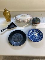 Photo showing six miscellaneous items on a table: yellow glass bottle, white covered dish with floral decoration, round lidded container, small cast iron skillet, black plate with skull design, and blue and white Fiesta plate.