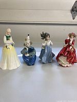 Four porcelain figurines displayed side by side on a white surface: Lladro girl with bird, Royal Doulton clown, female in blue dress, female in red dress.