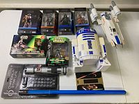 Overview of the lot showing six boxed Star Wars action figures, two spacecraft toys, R2-D2 figure, and a packaged lightsaber with blue blade and decals.