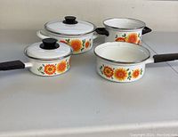 Four vintage enamel pots and pans arranged side-by-side showing white enamel with sunflower floral designs, black handles, and lids with black knobs.