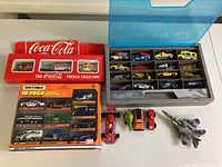 Collection showing Coca-Cola vehicle set, Matchbox starter pack in sealed packaging, small racing cars in a blue plastic box, and loose die cast vehicles and aircraft models on the table.
