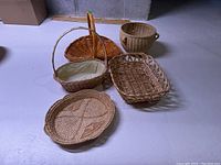 Five basketware items displayed on a floor: three wicker baskets, one woven grass tray, and one ceramic planter.