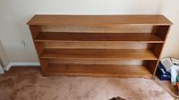 Full view of pine bookcase showing three shelves and wood grain finish against a wallpapered wall on carpeted floor.